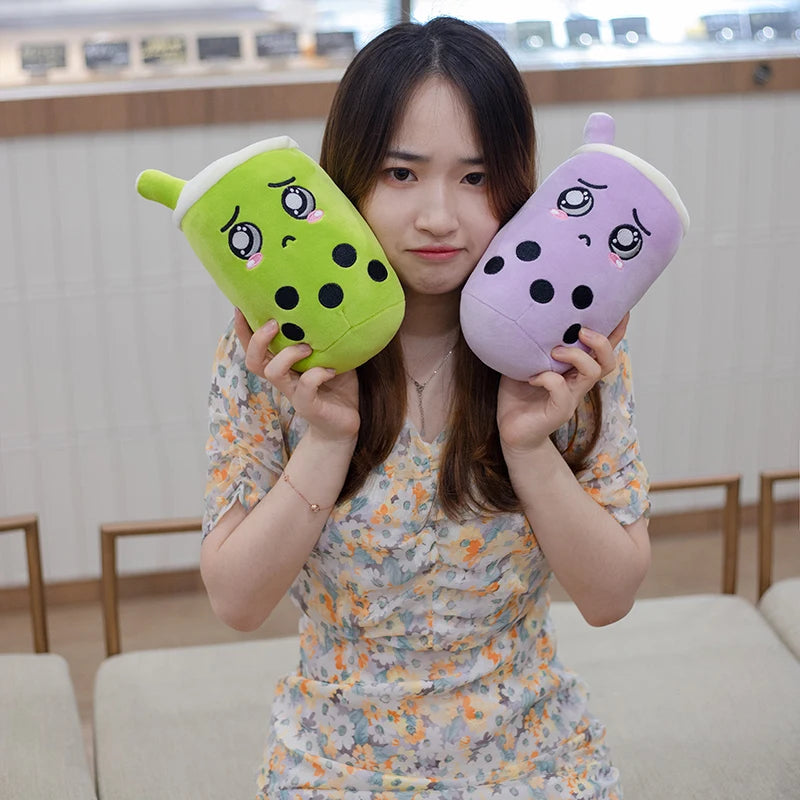 An image of a woman holding two crying boba tea plushies, one green matcha with tapioca, the other purple taro with tapioca. The woman is making a sour sad face, wearing a floral dress, and sitting on a cushion bench